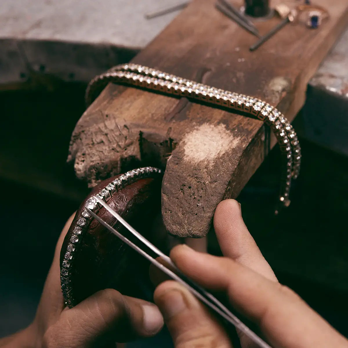 A close-up of a jeweler's hands using tweezers to place a diamond tennis bracelet onto a wooden workbench, with another band secured in a metal clamp above.