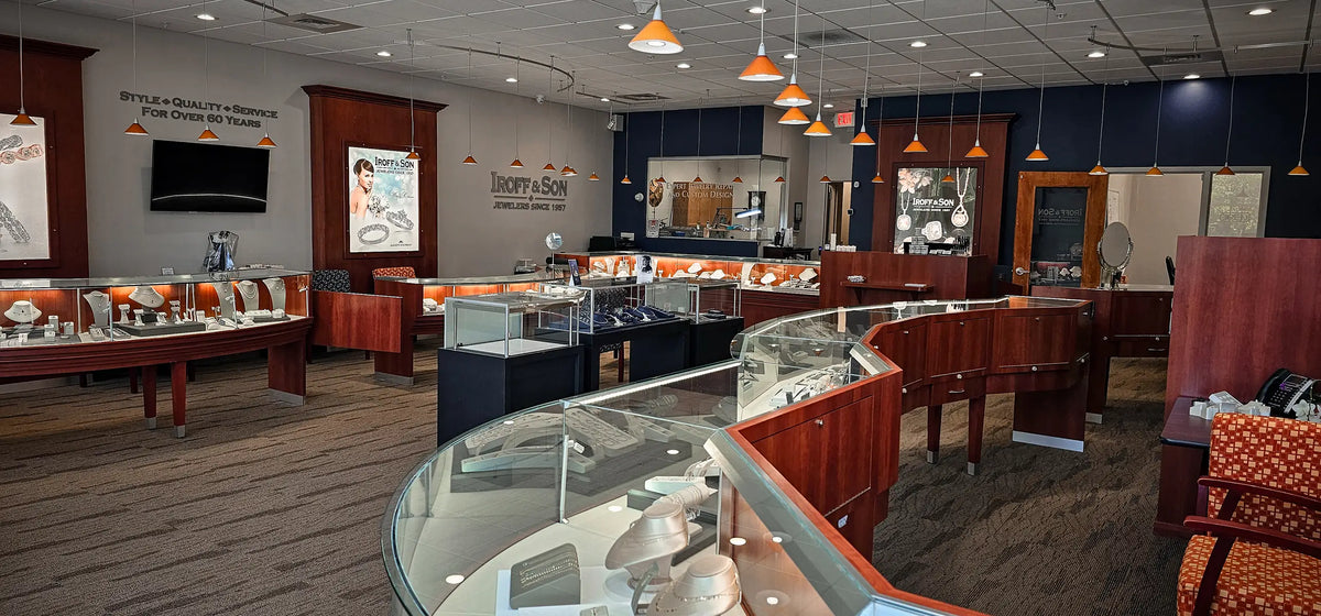 A wide interior view of the Iroff & Son jewelry showroom with multiple curved glass display cases, wood counters, hanging pendant lights, and framed wall art.
