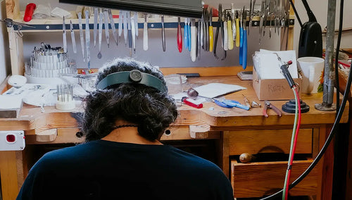 A jeweler sitting at a wooden workbench in a jewelry-making or craft workshop, wearing a dark headband with a magnifying lens. Tools and equipment hang on a rack above the workbench, and various crafting supplies are spread across the desk.