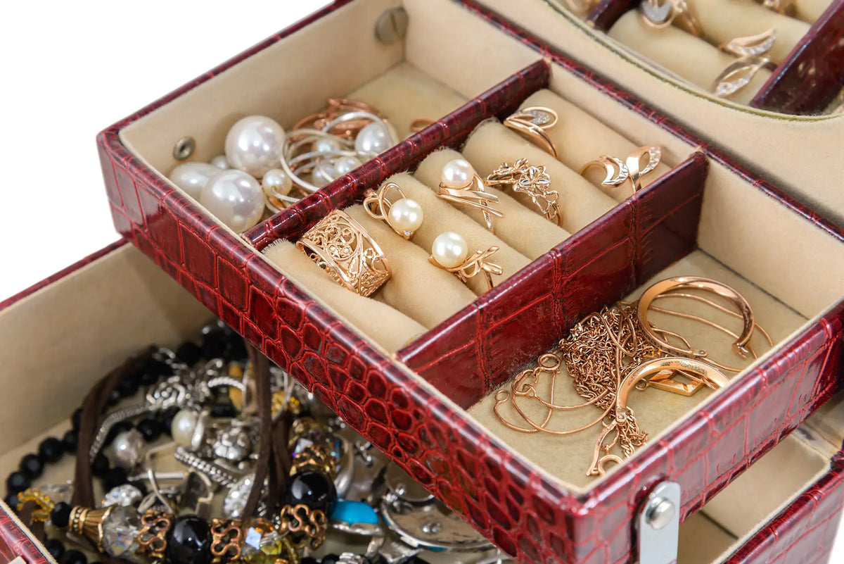 An open red crocodile-embossed jewelry organizer box with cream velvet compartments, filled with rings, earrings, and pearl jewelry, with more jewelry visible in the lower tray.